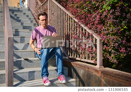 Young Man With Laptop Sitting on Outdoor Steps Working Remotely in Urban Setting 131906585