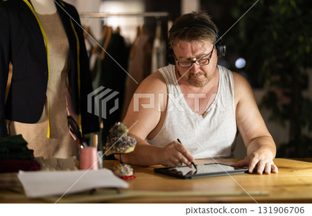 Middle-aged man wearing earphones drawing on tablet in sewing workshop at night 131906706