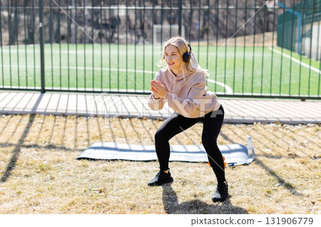 Young woman wearing headphones performing squats outdoors 131906779