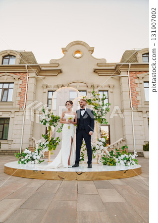 The bride and groom on a wedding arch decorated with greenery and white flowers. The bride and groom on a wedding arch decorated with greenery and white flowers. 131907002
