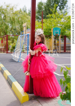 A cute little girl in an elegant lush pink dress is playing on the sports ground 131907134