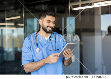 Young male doctor smiling, wearing scrubs and a stethoscope, interacting with a digital tablet and technology, managing healthcare data in a modern medical office setting 131907642