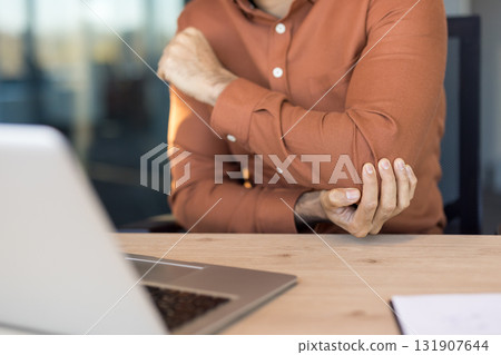 Man at office desk wincing while holding his elbow beside a laptop, showing joint inflammation and tendonitis from repetitive desk work and poor ergonomics 131907644