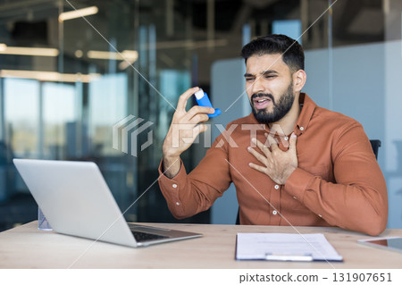 Young man experiencing an asthma attack while working at his office desk, holding an inhaler and clutching his chest, indicating discomfort and breathing difficulties Young man experiencing an asthma attack while working at his office desk, holding an inhaler and clutching his chest, indicating discomfort and breathing difficulties 131907651