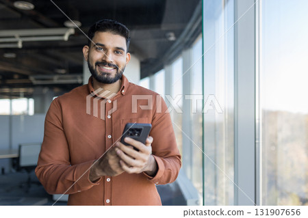 Indian businessman smiling at camera, holding a smartphone near a large window in a contemporary high-rise office, representing communication, technology, and success 131907656