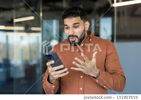 Young man standing in an office, holding a smartphone and expressing surprise, shock, and concern while receiving unexpected bad news or a surprising message 131907659