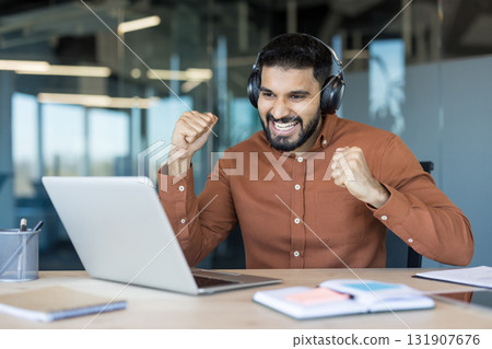 Young man wearing headphones happily celebrating business success or achieving a goal while looking at a laptop computer in a modern office workplace, fists pumping in excitement Young man wearing headphones happily celebrating business success or achieving a goal while looking at a laptop computer in a modern office workplace, fists pumping in excitement 131907676