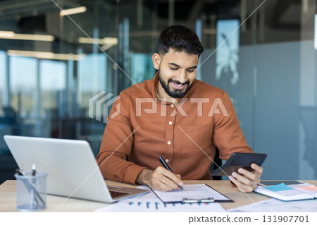 Indian businessman smiling at desk while reviewing paperwork with laptop and calculator, managing budgeting, taxes and finances in a modern office environment 131907701