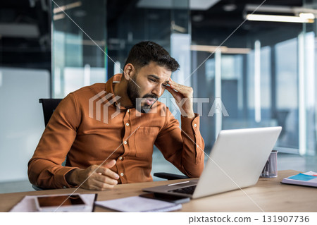 Young businessman sitting at his desk in a modern office, feeling overwhelmed and exhausted, rubbing his forehead with a pained expression while working on his laptop 131907736