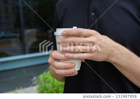 man holds disposable coffee cup with both hands and warms his hands on hot mug. coffee break. White paper kraft disposable cup for coffee with white plastic lid. Take-away cup. man holds disposable coffee cup with both hands and warms his hands on hot mug. coffee break. White paper kraft disposable cup for coffee with white plastic lid. Take-away cup. 131907958