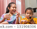 Two Asian Girls Eating Snacks and Using Smartphone Together at Home 131908306