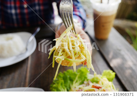 Close up hand holding fork with Thai spicy mango salad with crispy fish and vegetable in white dish on wooden table Close up hand holding fork with Thai spicy mango salad with crispy fish and vegetable in white dish on wooden table 131908371