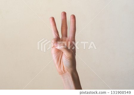 A hand with a light skin tone is raised, showing the number three with fingers extended. The background is a neutral color, emphasizing the gesture. A hand with a light skin tone is raised, showing the number three with fingers extended. The background is a neutral color, emphasizing the gesture. 131908549
