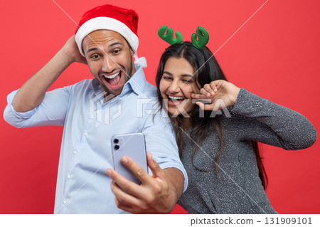 Merry young fun couple wearing Santa hat and deer headband doing selfie shot on mobile cell phone Merry young fun couple wearing Santa hat and deer headband doing selfie shot on mobile cell phone 131909101