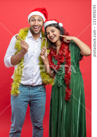 Happy couple dressed in Santa hats and tinsel celebrating new year together holding candy canes 131909295