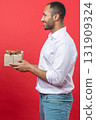 Smiling man in white shirt holding present box with gift ribbon bow isolated over red background 131909324