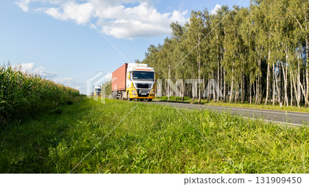 Two Yellow Trucks Driving on Highway Through Forest Landscape and Field 131909450