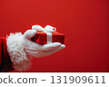 Front view of Santa Claus hands is holding red gift box with golden ribbon bow. Red background. Merry Christmas. Flat lay. Santa with present box. Wide long banner, Panoramic. Copy space. Giftbox 131909611