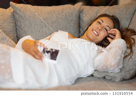 Pregnant woman in white dress smiling and holding ultrasound photo 131910554