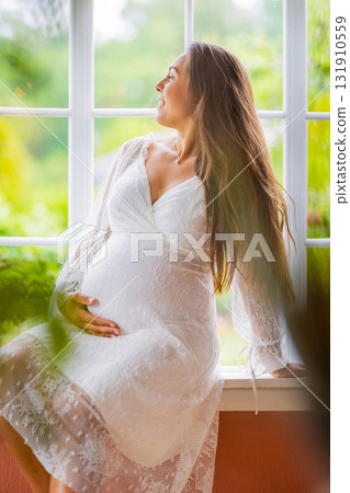 Pregnant woman in a white dress enjoying serene outdoor sunlight 131910559