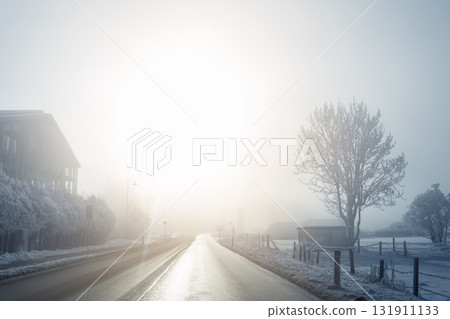 Scenic quiet winter road frosty valley covered mist fog and snow. Bare trees and distant mountains glow softly under the pale morning light. Alpine mountain village winter morning sunrise Scenic quiet winter road frosty valley covered mist fog and snow. Bare trees and distant mountains glow softly under the pale morning light. Alpine mountain village winter morning sunrise 131911133