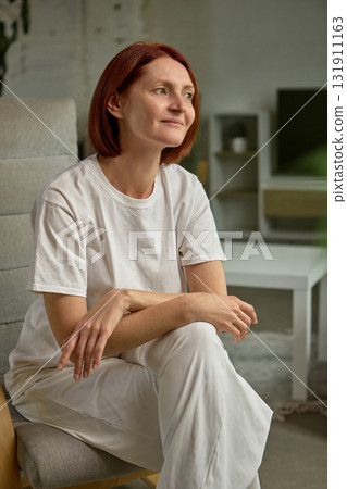 Relaxed woman with short red hair sitting on chair at home in soft daylight Relaxed woman with short red hair sitting on chair at home in soft daylight 131911163