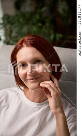Portrait of calm red-haired woman smiling softly while relaxing indoors in natural light 131911177