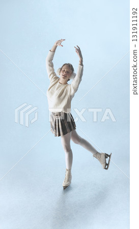 Smiling female skater performing joyful pose on ice in cozy winter outfit under soft light 131911192
