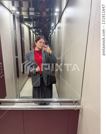 A woman taking a mirror selfie inside an elevator in stylish layered outfit. Urban femininity, confidence and personal identity in reflective modern environment. A woman taking a mirror selfie inside an elevator in stylish layered outfit. Urban femininity, confidence and personal identity in reflective modern environment. 131911647