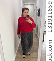 A woman standing in a hallway wearing a red sweater and metallic skirt. Everyday mood, quiet confidence and casual indoor atmosphere in modern home interior. 131911659