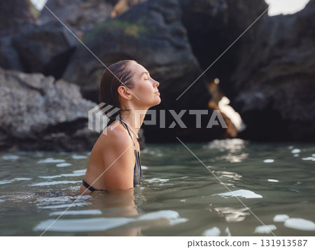 Young woman enjoying serene evening at sea in Krabi, Thailand during sunset. Calm waves, golden light, and peaceful atmosphere highlight tropical beauty and connection with nature. 131913587