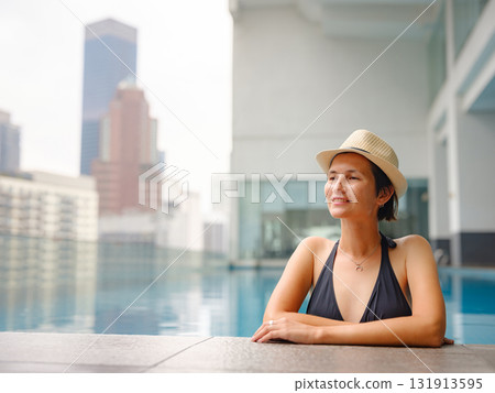 Young asian woman relaxing by pool at Kuala Lumpur hotel with view of surrounding skyscrapers, enjoying leisure time in vibrant urban setting. Young asian woman relaxing by pool at Kuala Lumpur hotel with view of surrounding skyscrapers, enjoying leisure time in vibrant urban setting. 131913595
