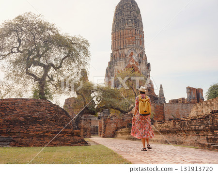 Young female tourist in dress exploring historic park of Ayutthaya, Thailand. Surrounded by ancient temples and statues, she enjoys peaceful atmosphere and rich cultural heritage. 131913720