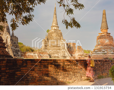 Young female tourist in dress exploring historic park of Ayutthaya, Thailand. Wat Phra Si Sanphet ancient temples and statues, she enjoys peaceful atmosphere and rich cultural heritage. 131913734