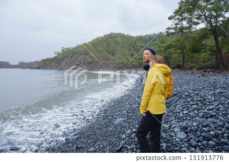 Couple enjoys Turkish coastline. Sitting on stones, watching sea waves, relaxing together, slow romantic moment near calm blue water under soft winter light. 131913776