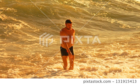 Man Ocean Sunset: Bare-chested man stands in foamy golden waves at beach during beautiful golden hour. Man Ocean Sunset: Bare-chested man stands in foamy golden waves at beach during beautiful golden hour. 131915413