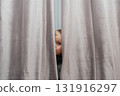 Small child peeks out from behind drapery, curious expression caught in indoor natural light 131916297