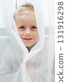 Cheerful boy smiles as he peeks from behind a sheer white curtain bright indoor portrait with playful mood 131916298