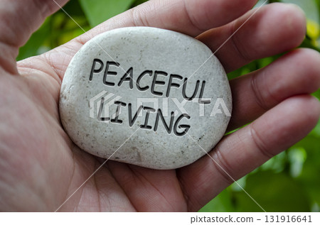 Hand holding a white stone with PEACEFUL LIVING text engraved on it, expressing life purpose of calmness and emotional stability Hand holding a white stone with PEACEFUL LIVING text engraved on it, expressing life purpose of calmness and emotional stability 131916641