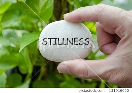 Hand holding a white stone with STILLNESS text engraved on it, representing life purpose of tranquility and peace of mind 131916660