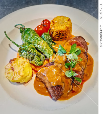 A vibrant dish of sliced beef steak topped with rich orange sauce and garnished with fresh microgreens, fire-roasted corn, grilled padron peppers, charred cherry tomatoes and cheesy potato gratin 131916704