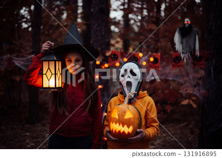 Two children in halloween costumes in the woods. Autumn holiday decoration, jack-o-lantern, creepy mask, witch hat, ghost on the background. 131916895
