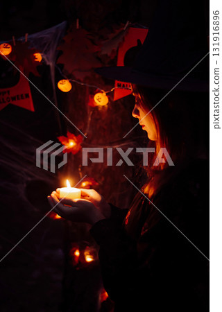 Young woman in the woods at night holding a candle. Wearing black dress, witch hat, halloween mystery decorations around. Young woman in the woods at night holding a candle. Wearing black dress, witch hat, halloween mystery decorations around. 131916896