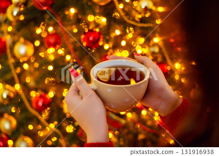 A cup of tea with lemon in hands of young girl, beautiful warm lights of christmas tree on the background. New year, christmas, cozy festive magical atmosphere at home A cup of tea with lemon in hands of young girl, beautiful warm lights of christmas tree on the background. New year, christmas, cozy festive magical atmosphere at home 131916938