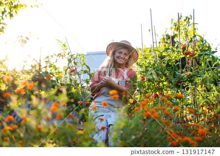 Older female farmer resting among flowers in garden, having cup of herbal tea. 131917147