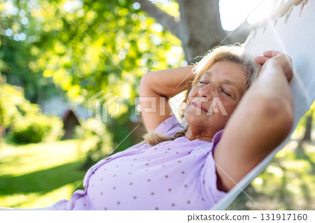 Senior woman lying in a hammock with closed eyes, quiet time in garden. 131917160