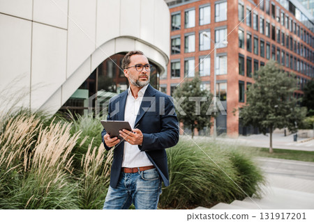 Businessman in front of office building, holding tablet and reading something. 131917201