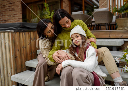 Mother, grandmother and granddaugter sitting on wooden patio, hugging. 131917361