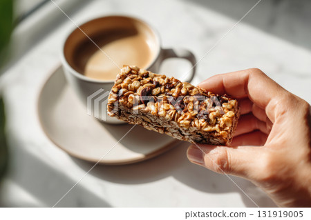 Hand holding granola energy bar above cup of coffee on bright table Hand holding granola energy bar above cup of coffee on bright table 131919005