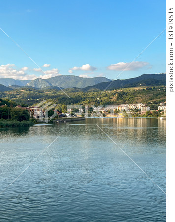 A small coastal town with houses and buildings, located by a calm river, surrounded by wooded hills A small coastal town with houses and buildings, located by a calm river, surrounded by wooded hills 131919515